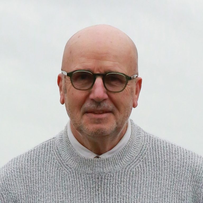 Homme âgé avec des lunettes, portant un pull noir, fond flou de ciel nuageux.