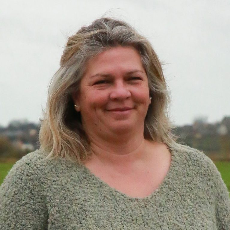 Femme souriante avec des cheveux longs et blonds, au bord d'un champ verdoyant.
