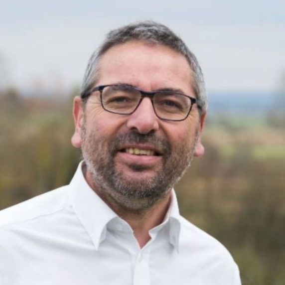 Homme souriant avec des lunettes, portant une chemise bleue, en arrière-plan naturel.